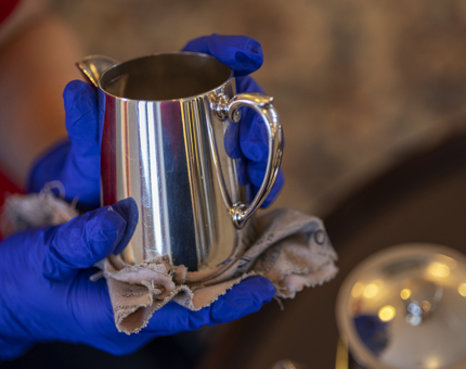 A close up of hands wearing blue gloves polishing a small silver jug. 