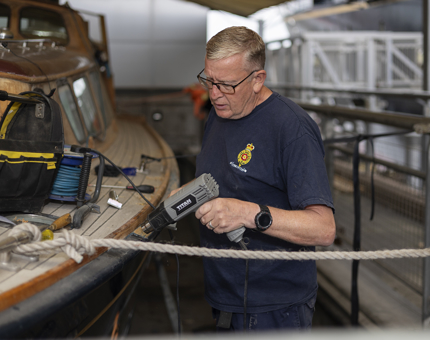 A Maintenance man holding a heat gun, removing varnish off of the Royal Barge. 