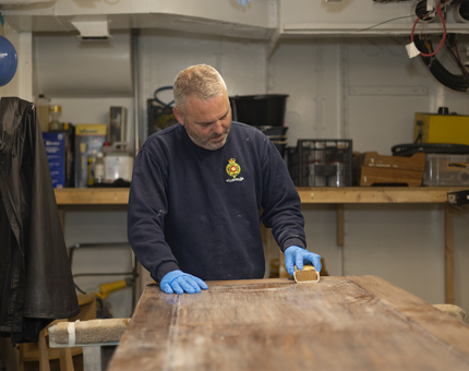 A Maintenance man is holding sandpaper, sanding down a wooden door. 