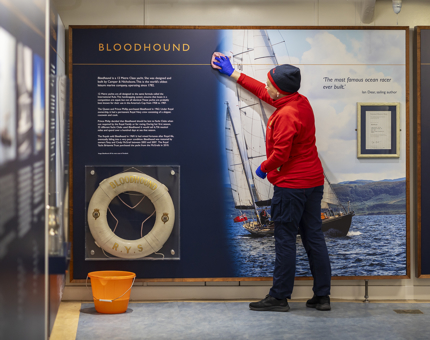 A Housekeeper wearing a red fleece is wiping a display board in the Sailing exhibition.