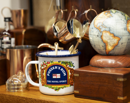 An enamel mug decorated with Pusser Rum design. There are brass mugs and a small globe in the background. 