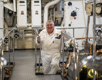 A man wearing white overalls climbing down a ladder in the Engine Room. 