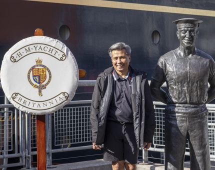 A Man poses for a photo next to a statue of Norrie, the longest serving Royal Yachtsmen. There is a perry buoy next to them with the Britannia crest on it. 