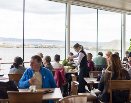 In the Royal Deck Tearoom visitors sit at the tables. A server is placing plates of food on a table by the windows where you can see the views of the Port of Leith.