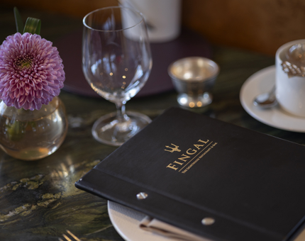 In The Lighthouse Restaurant, a table is set for Afternoon Tea. There is a small round vase with a purple flower, a menu in a black cover sits on a white plate. 
