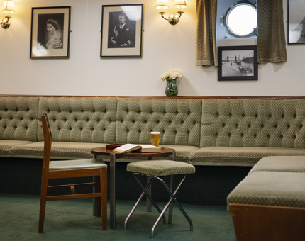 In the Chief Petty Officers Mess, the benches are covered in green fabric. There is a small tables with a chair and a stool next to it. In the background there are pictures of the Royal Family hanging on the wall. 