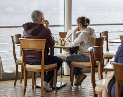 Two visitors are sitting at a table in the Tearoom, they are facing the windows where there is a view of the water and port.