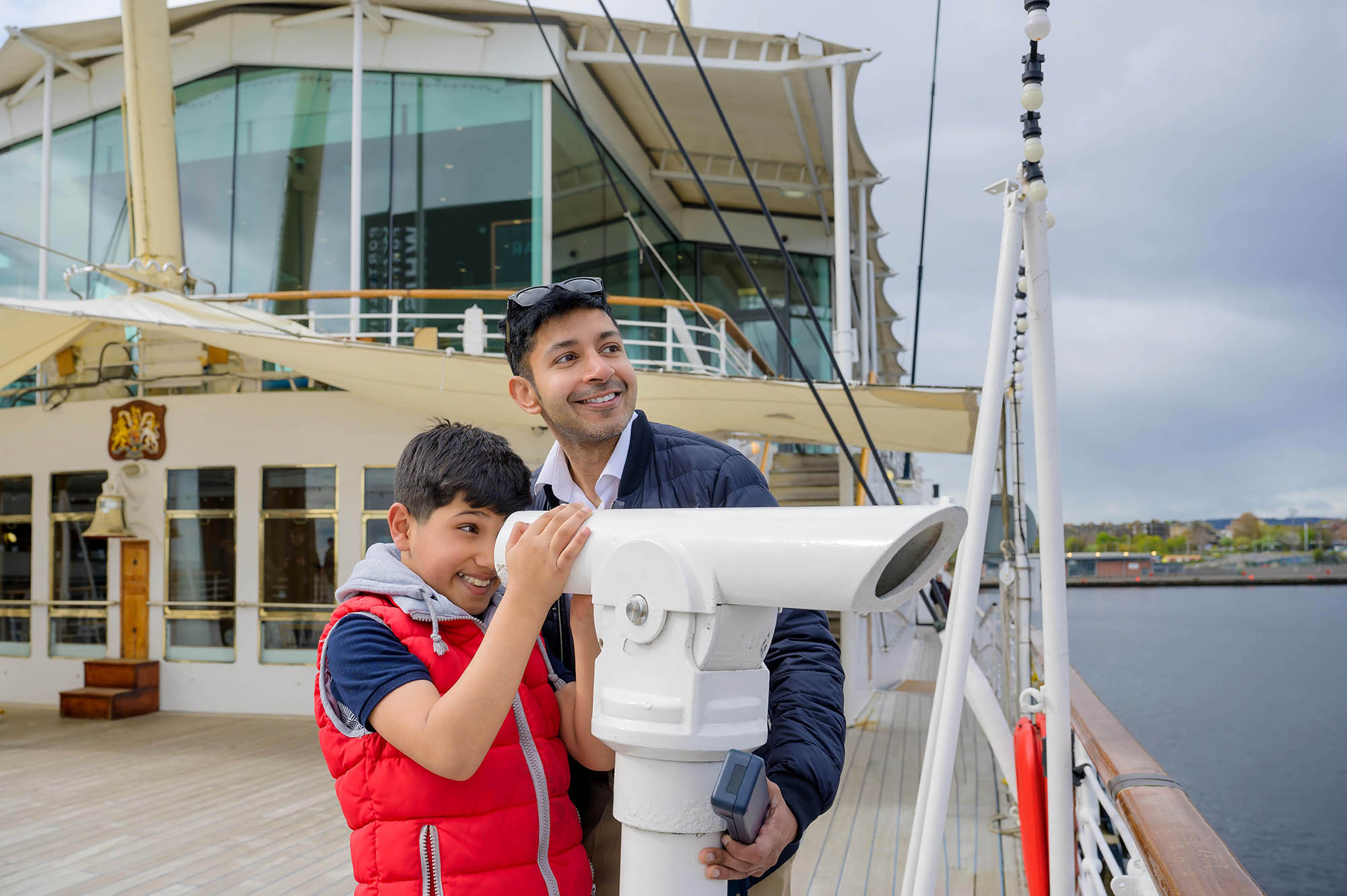 A man and boy on The Royal Yacht Britannia's Verandah Deck in Edinburgh looking through a telescope.