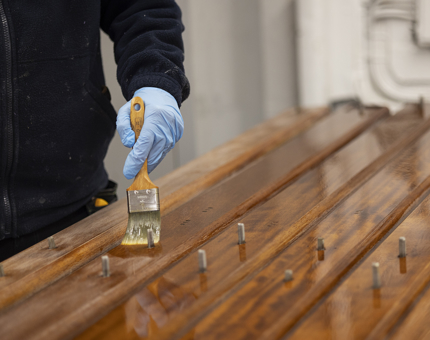 A close-up of a hand wearing a blue glove, holding a paint brush, varnishing handrails. 