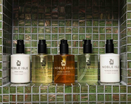 Five bottles of Noble Isle bath and body products sitting on a green tiled shelf in a cabin's bathroom aboard Fingal Hotel.