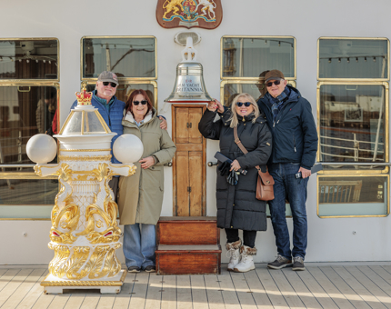 Four people pose for a photo by Britannia's Bell and binnacle on the Verandah Deck. 