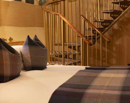 Aboard Fingal in Leith, a  close up of a bed with a tartan blanket and pillows is in the foreground, a spiral staircase is in the background.