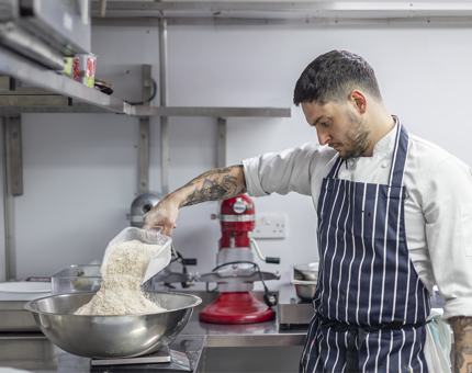 A chef pouring flour into a metal bowl to weigh it. 