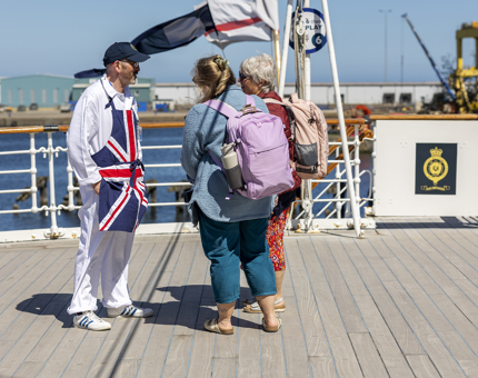 A man wearing white overalls and a union jack apron talking to to two women on the Verandah Deck. You can see the water behind them. 