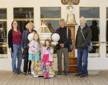 A Family with two women, two men and two girls, posing for a photo by Britannia's Bell and binnacle. 