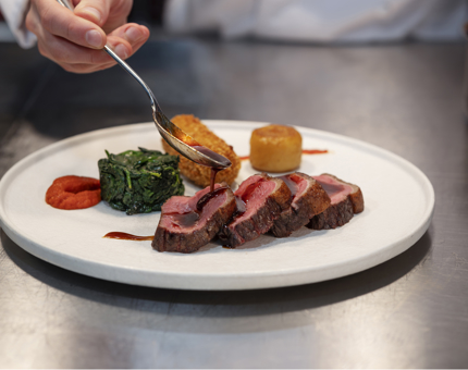 A hand holding a spoon, placing sauce on a plate of beef and vegetables. 