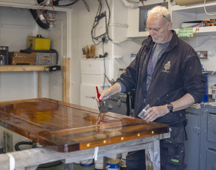 In the workshop, a Maintenance man stands by a wooden door holding a pot of varnish. He is applying a coat of varnish with a paintbrush. 
