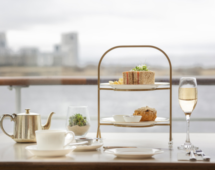 A tea stand containing sandwiches and scones with a silver teapot and glass of fizz on a table in the Royal Deck Tearoom. There is a waterside view behind. 