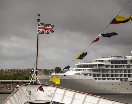 A view of the bow of the ship. There is a Union Jack flag flying from it and a line of dress flags in the foreground. In the background there is a large cruise ship called the Silver Endeavour.